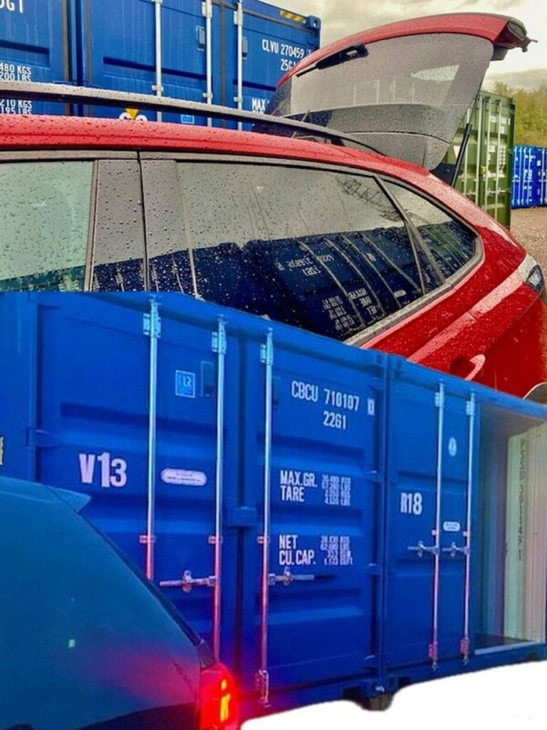 Red car with open boot next to stacked blue shipping containers.