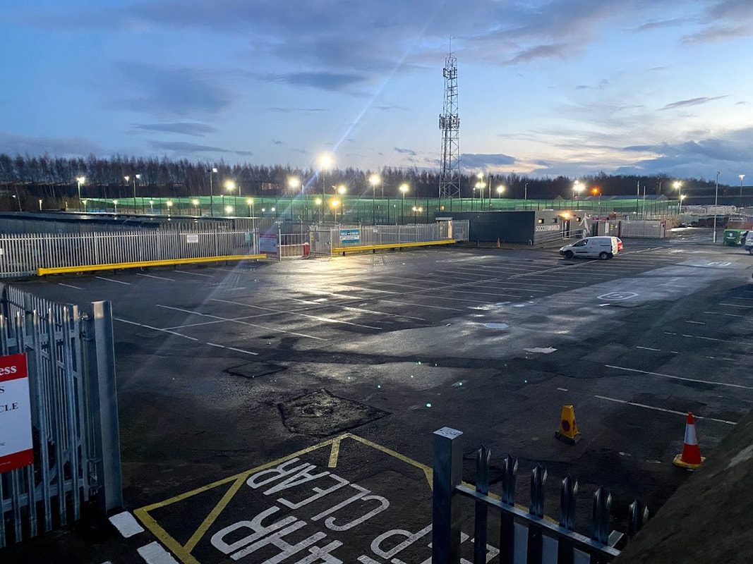 Empty parking area at dusk, with illuminated sections and a distant tower at Rome Street Depot Carlisle.