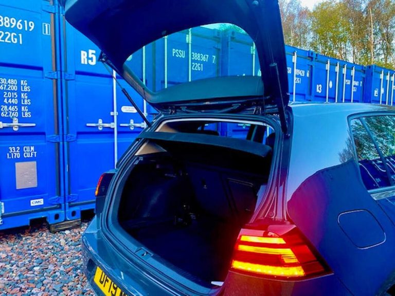 Open boot of a blue car parked near blue shipping containers.