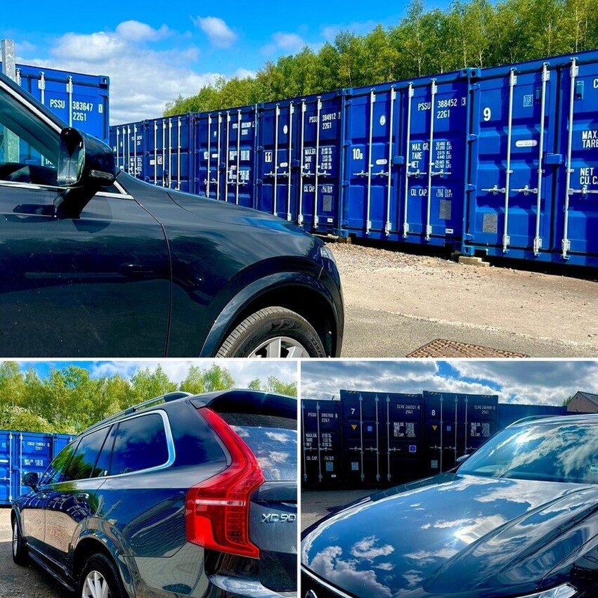 Parked black car in front of a row of blue shipping containers against a clear sky.